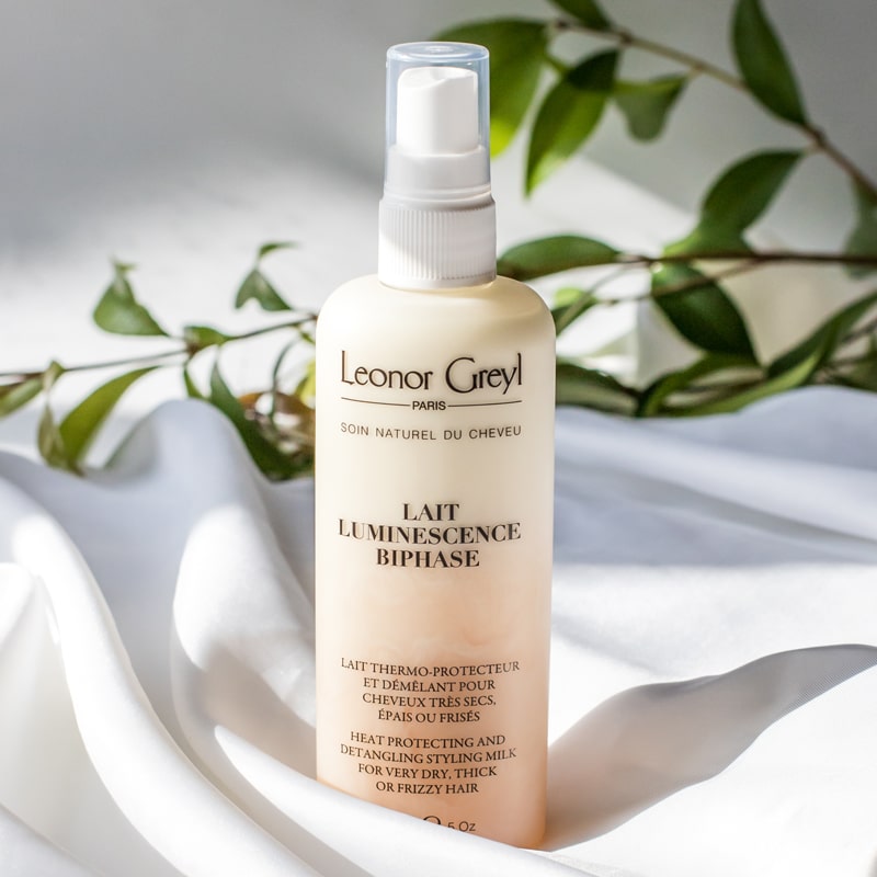 Lifestyle shot of Leonor Greyl Lait Luminescence Bi-Phase (150 ml) with white fabric and branch with leaves in the background