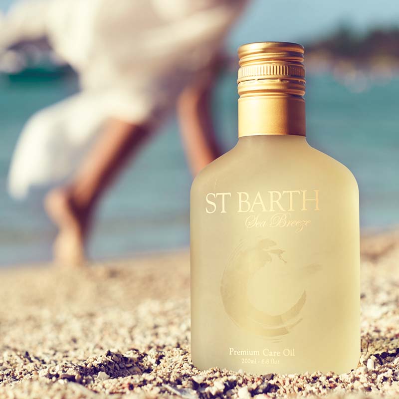 Lifestyle shot of Ligne St. Barth Premium Care Oil 6.8 oz in the sand with woman walking along the beach in the background