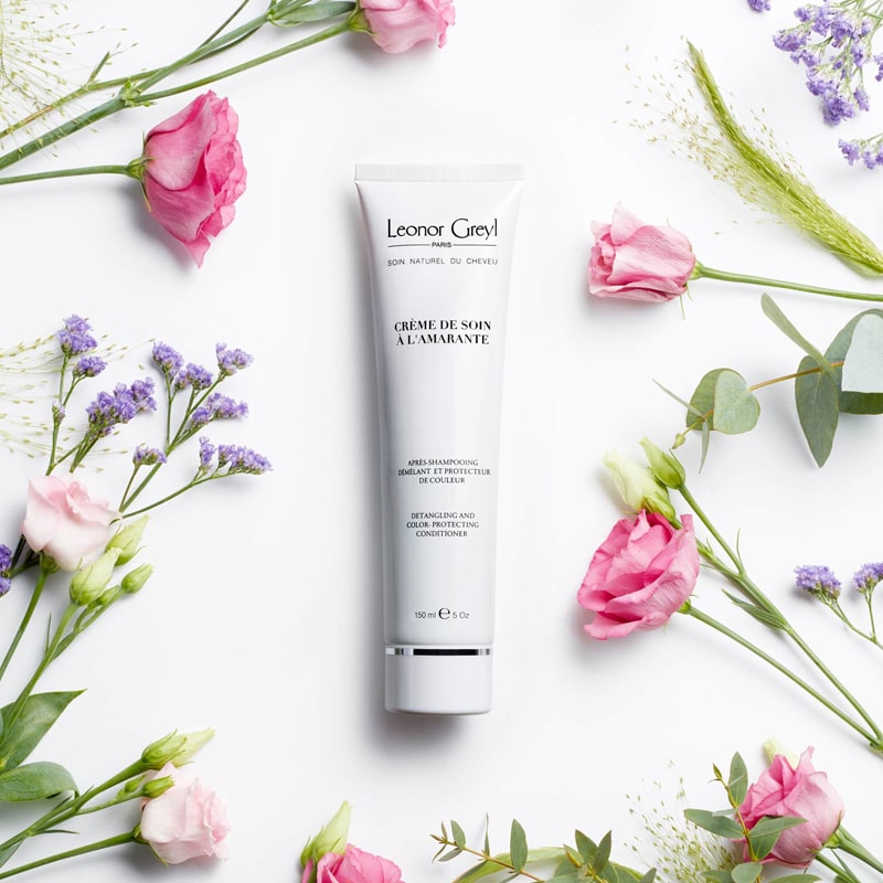 Lifestyle shot top view of Leonor Greyl Creme de Soin a L&#39;Amarante (150 ml) with pink roses, purple flowers and leaves in the background