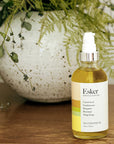 Esker Beauty Restorative Oil (4 oz) shown on wood table with vase in the background with green fern leaves and ivy