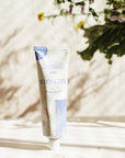 Lifestyle shot of Minois Paris Creme Douce (Gentle Cream) (100 ml) with vase of flowers in the background