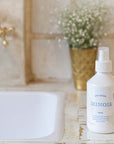 Lifestyle shot of Minois Paris Eau Legere (Light Water) (250 ml) on sink vanity with vase of white flowers in the background