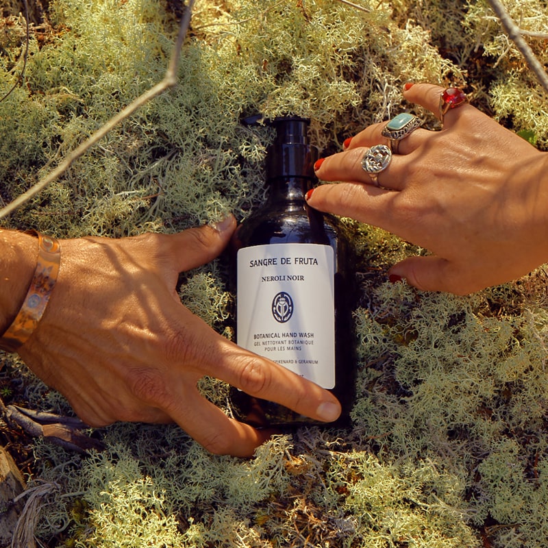 Lifestyle shot of Sangre de Fruta Botanical Hand Wash Neroli Noir (500 ml) top view with moss in the background and man an woman&#39;s hand reaching for bottle 
