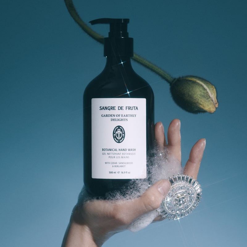 Lifestyle shot of Sangre de Fruta Botanical Hand Wash Garden of Earthly Delights (500 ml) in palm of model with soap suds and flower bud in the background