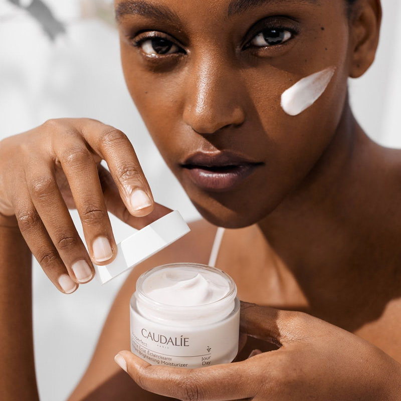 Caudalie Vinoperfect Instant Brightening Moisturizer shown on model&#39;s face and in her hand an open jar