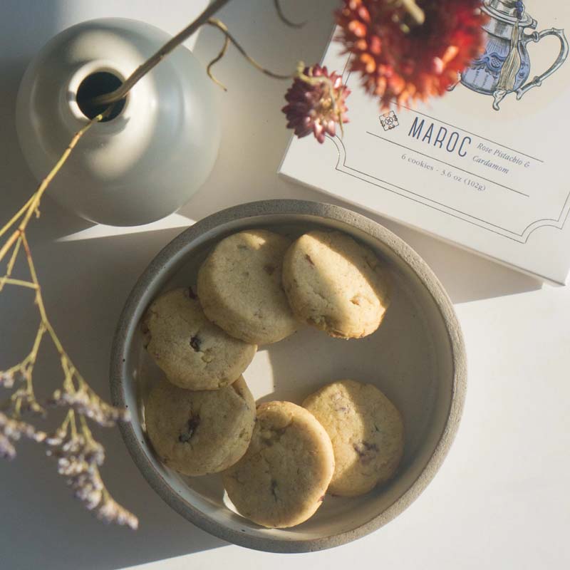 Lifestyle shot of Makabi &amp; Sons Rose Pistachio Cardamom Cookies - Maroc, 6 cookies shown in a dish with box and vase with flowers in the background