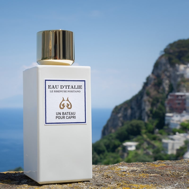 Lifestyle shot of Eau d&#39;Italie Un Bateau Pour Capri Eau de Parfum Spray (100 ml) with Positano hillside in the background