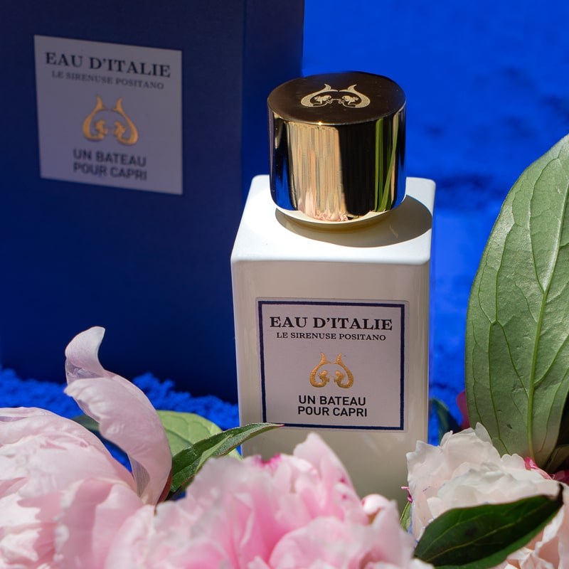 Lifestyle shot of Eau d&#39;Italie Un Bateau Pour Capri Eau de Parfum Spray (100 ml) with box and pink flowers and green leaves in the foreground