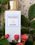Lifestyle shot of Eau d'Italie Rosa Greta Eau de Parfum Spray bottle (100 ml) with pink rose buds and leaves in the background