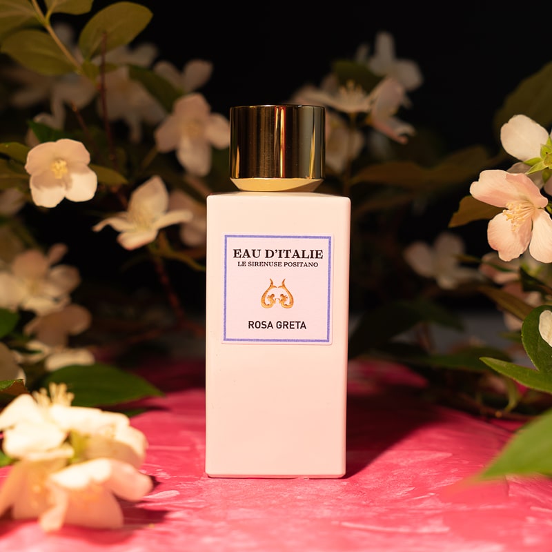 Lifestyle shot of Eau d&#39;Italie Rosa Greta Eau de Parfum Spray bottle (100 ml) on pink textured fabric and white flowers in the background