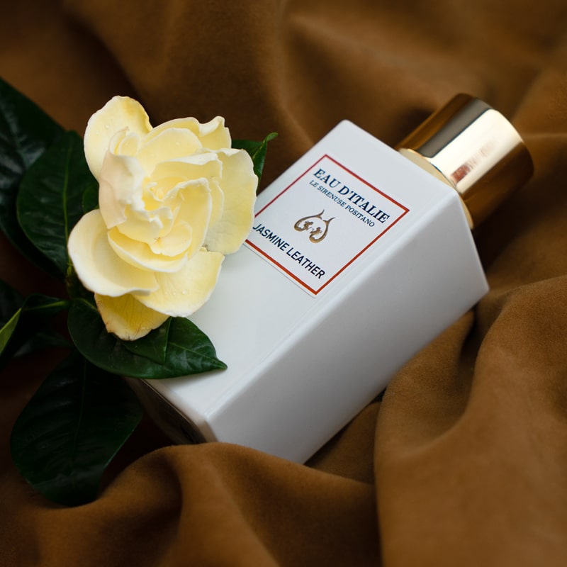 Lifestyle shot of Eau d'Italie Jasmine Leather Eau de Parfum Spray (100 ml) on brown fabric with jasmine flower