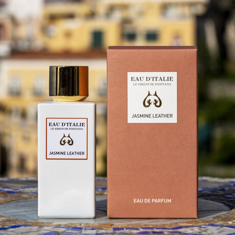 Lifestyle shot of Eau d'Italie Jasmine Leather Eau de Parfum Spray (100 ml) with box on tiled mosaic table and the Positano hillside in the background