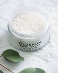 Olverum Bath Salts (200 g) shown top view with lid off and bath salts and leaves in the background