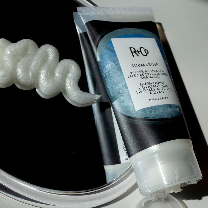 R+Co Submarine Water Activated Enzyme Exfoliating Shampoo showing a squiggle of the shampoo on a mirror beside tube