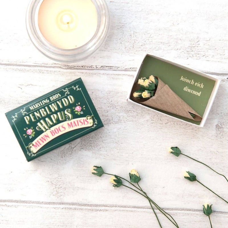 Lifestyle shot of Marvling Bros Ltd Welsh Birthday Folk Art Mini Bouquet In A Matchbox with flowers out of bouquet and next to a candle