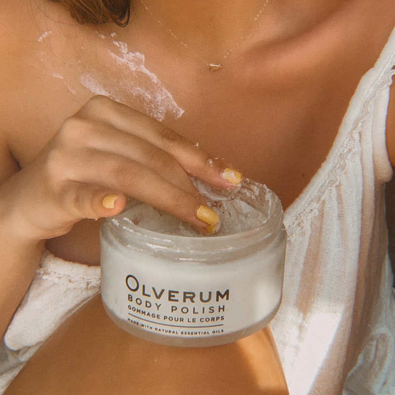 Olverum Body Polish shown with model taking more product and some applied to collarbone area