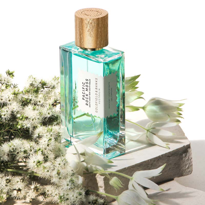 Lifestyle shot of Goldfield &amp; Banks Pacific Rock Moss Perfume 100 ml on stone slab with white flowers in the background and foreground