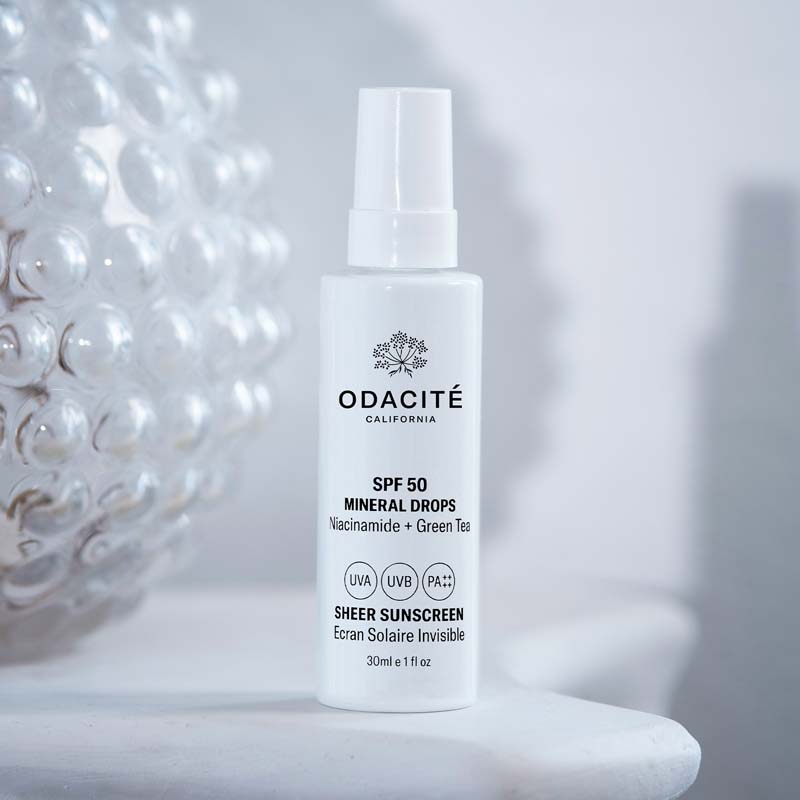 Lifestyle shot of Odacite SPF 50 Mineral Drops Sheer Sunscreen (30 ml) sitting on white table and clear glass globe in the background
