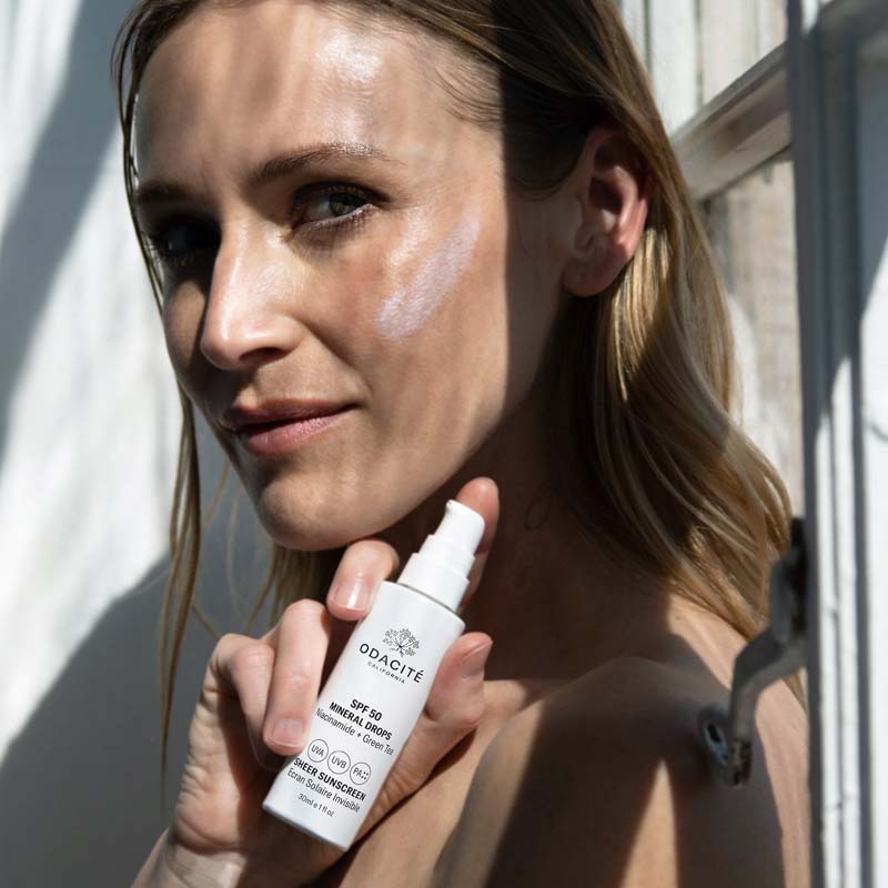 Close up of model holding Odacite SPF 50 Mineral Drops Sheer Sunscreen (30 ml) with smear applied to cheek