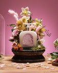 Santa Maria Novella Rosa Gardenia Fluid Body Cream sitting on a stand with a floral arrangement