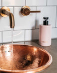 NEOM Organics Real Luxury Ceramic Hand Wash Dispenser & Refill showing on a counter next to a running faucet and copper bowl