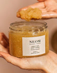 Lifestyle shot of Neom Organics Great Day Body Scrub with model scooping out with hand scrub