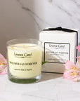 Lifestyle shot of Leonor Greyl Magnolias Forever Candle (11 oz) with box and pink flowers in the foreground and background