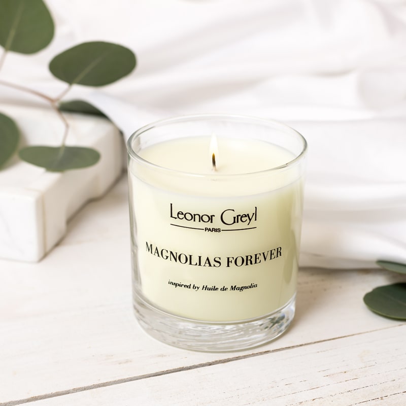 Lifestyle shot of Leonor Greyl Magnolias Forever Candle (11 oz) with green leaves in the background