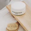 Susanne Kaufmann Body Butter - lifestyle shot showing jar on a board above a wooden handled brush