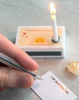 Marvling Bros Ltd Hello Sunshine Mindfulness Gift In A Matchbox showing lit candle in box and hand writing a note by the Citrine stone.