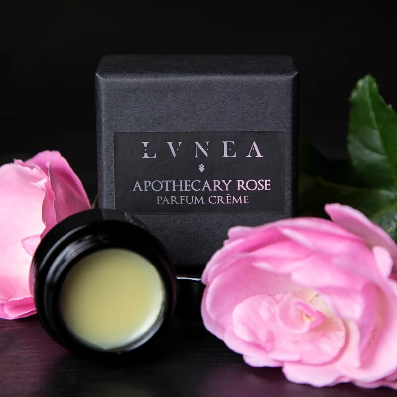Lifestyle shot of Lvnea Perfume Apothecary Rose Parfum Creme (10 g) with jar open and box and pink roses in the background and foreground