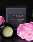 Lifestyle shot of Lvnea Perfume Apothecary Rose Parfum Creme (10 g) with jar open and box and pink roses in the background and foreground