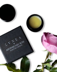 Lifestyle shot of Lvnea Perfume Apothecary Rose Parfum Creme (10 g) with jar open and box and pink rose in the foreground